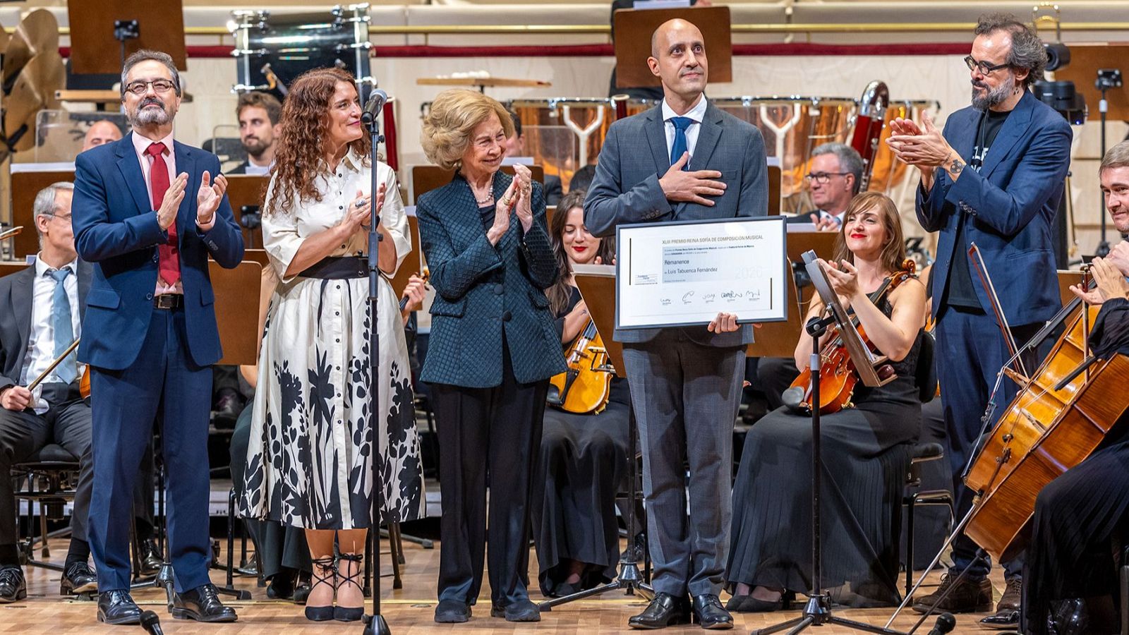 La Reina Doña Sofía aplaude al compositor Luis Tabuenca, quien sostiene un diploma enmarcado sobre el escenario de una sala de conciertos ante la presencia de la orquesta.