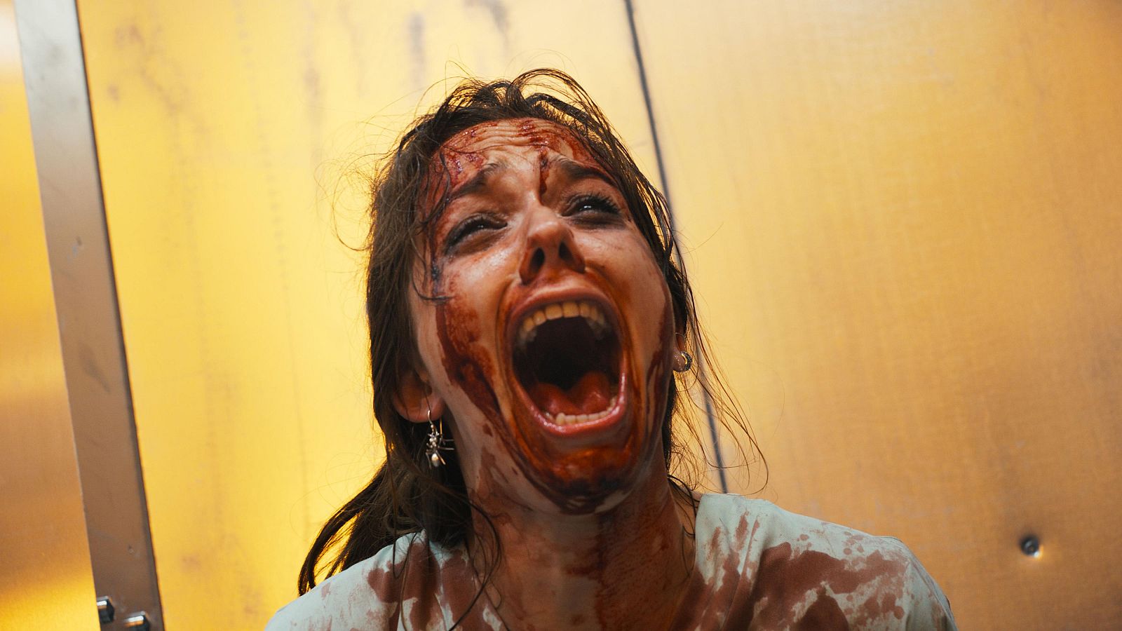 Mujer joven con expresión de pánico y rostro cubierto de sangre en un escenario industrial con paneles dorados.