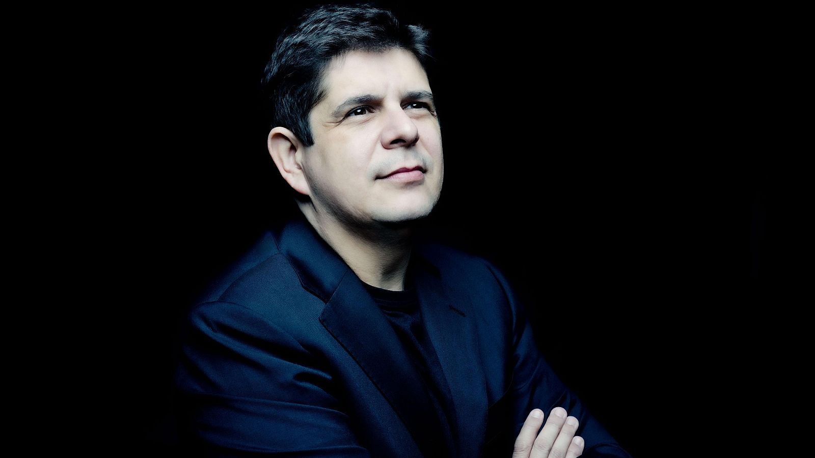 Retrato de medio cuerpo del pianista Javier Perianes con chaqueta azul marino y brazos cruzados sobre fondo negro.