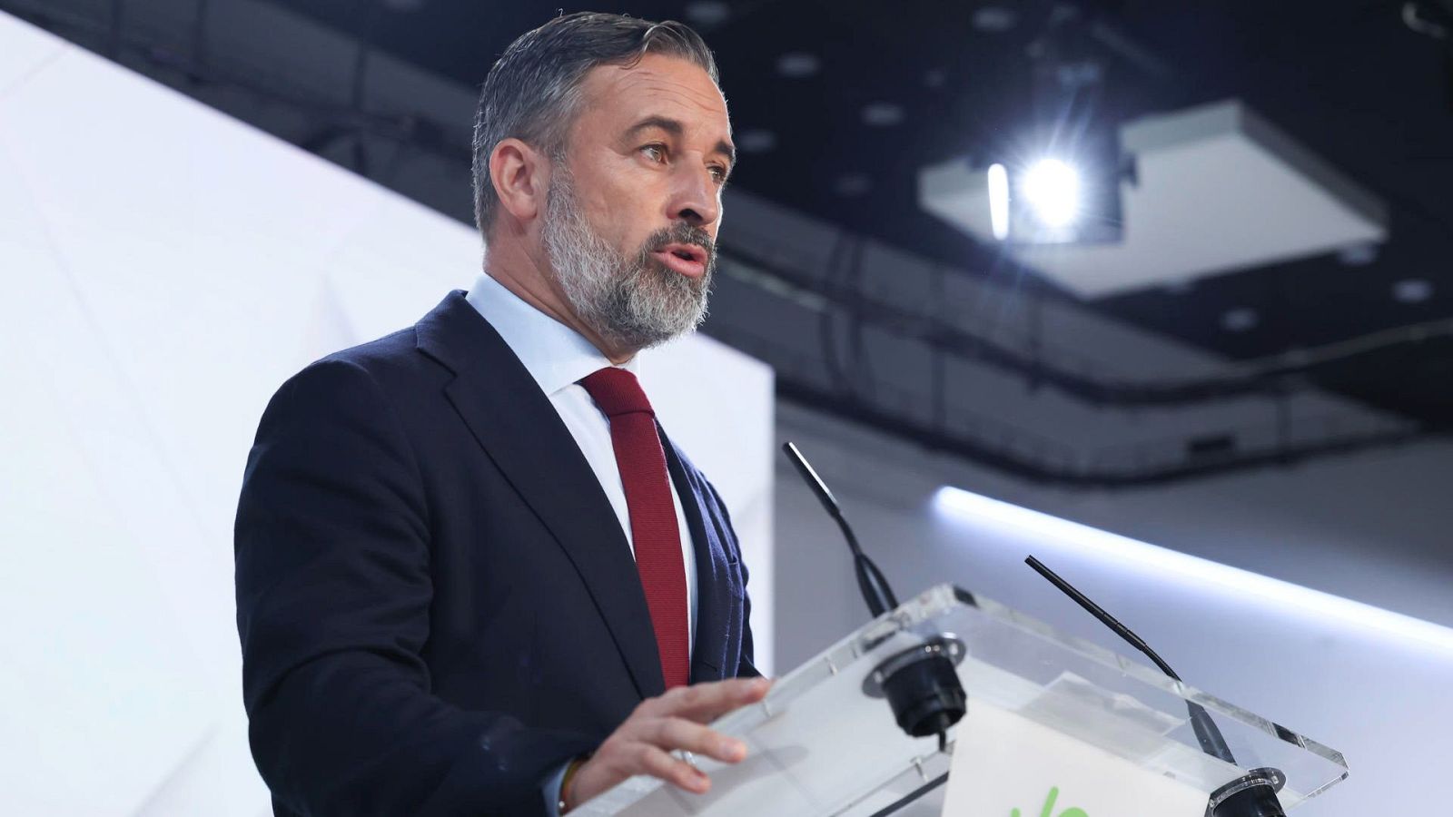 Santiago Abascal interviene tras un atril transparente con traje azul y corbata roja, con gesto serio y concentrado.