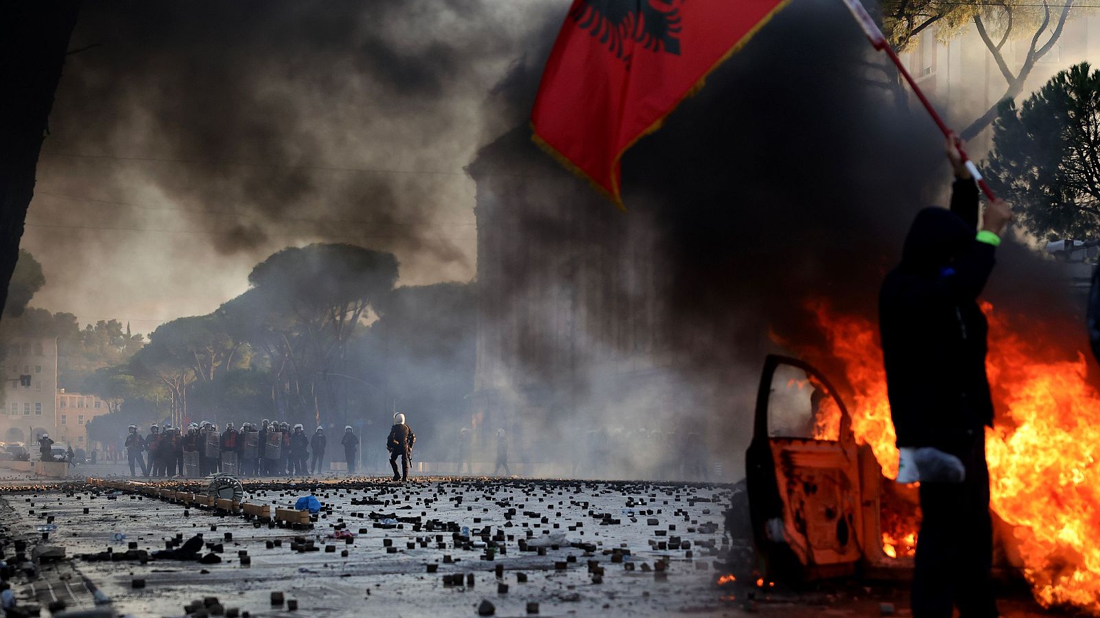 Un manifestante alza una bandera albanesa frente a un coche en llamas durante una protesta contra el Gobierno ante la sede de la oficina del primer ministro, en Tirana