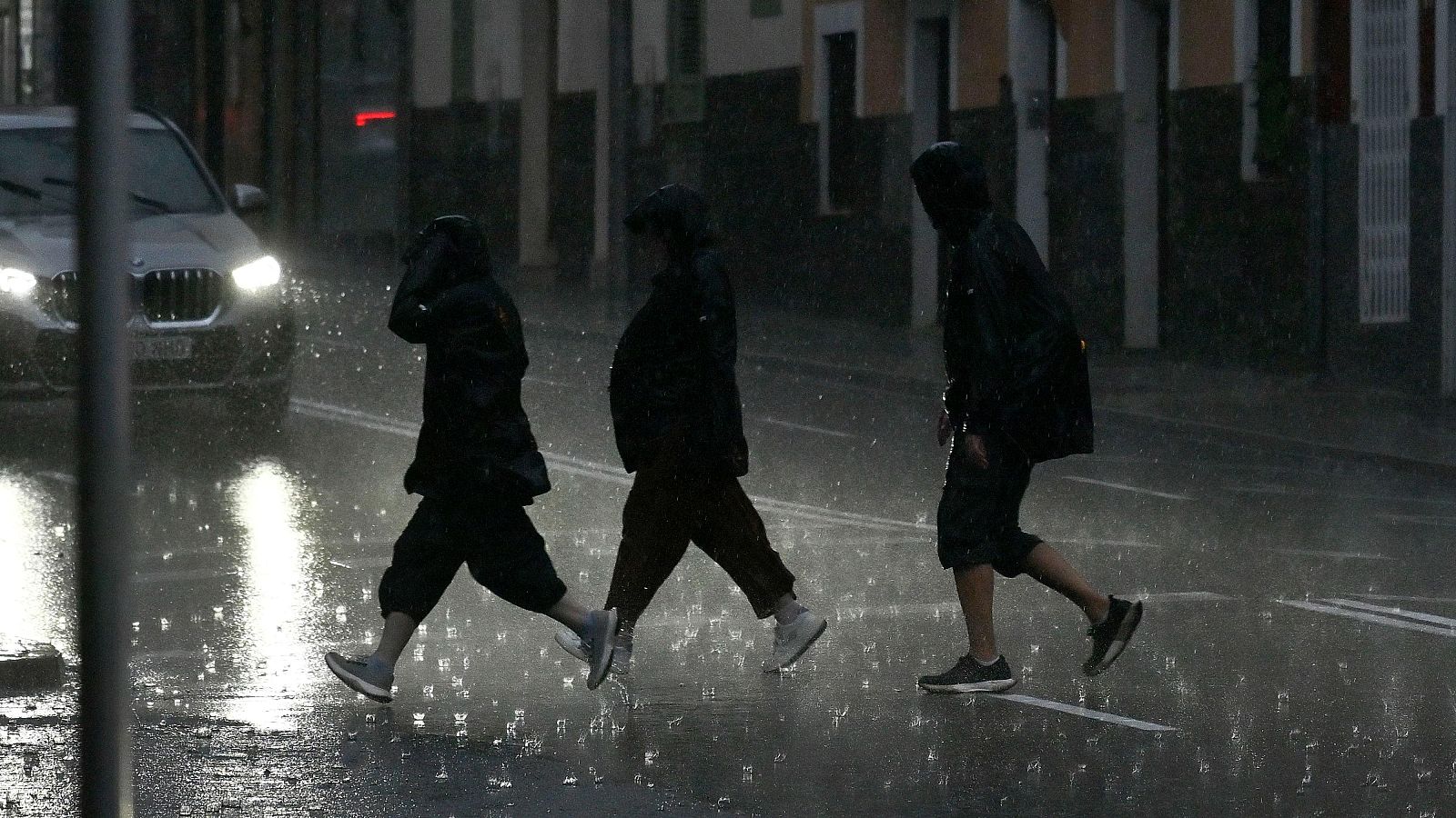 Tres persones creuen un carrer sota una pluja torrencial amb el terra enxangat.
