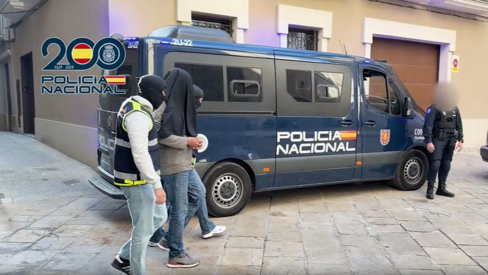Agents de la Policia Nacional custodien un detingut amb el rostre tapat durant una operació a la via pública, amb un furgó policial de fons.