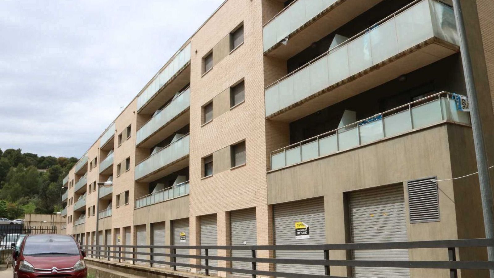 Edifici residencial de maó vist amb terrasses de vidre, planta baixa amb garatges i un cotxe vermell aparcat davant sota un cel ennuvolat.