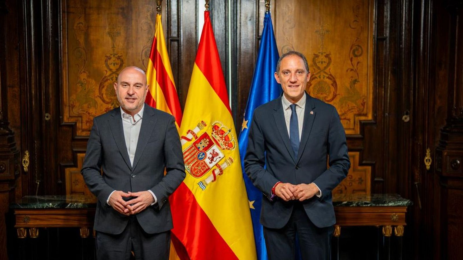 Carlos Prieto i José Crespín posen formalment en una sala oficial davant de les banderes de Catalunya, Espanya i la Unió Europea.