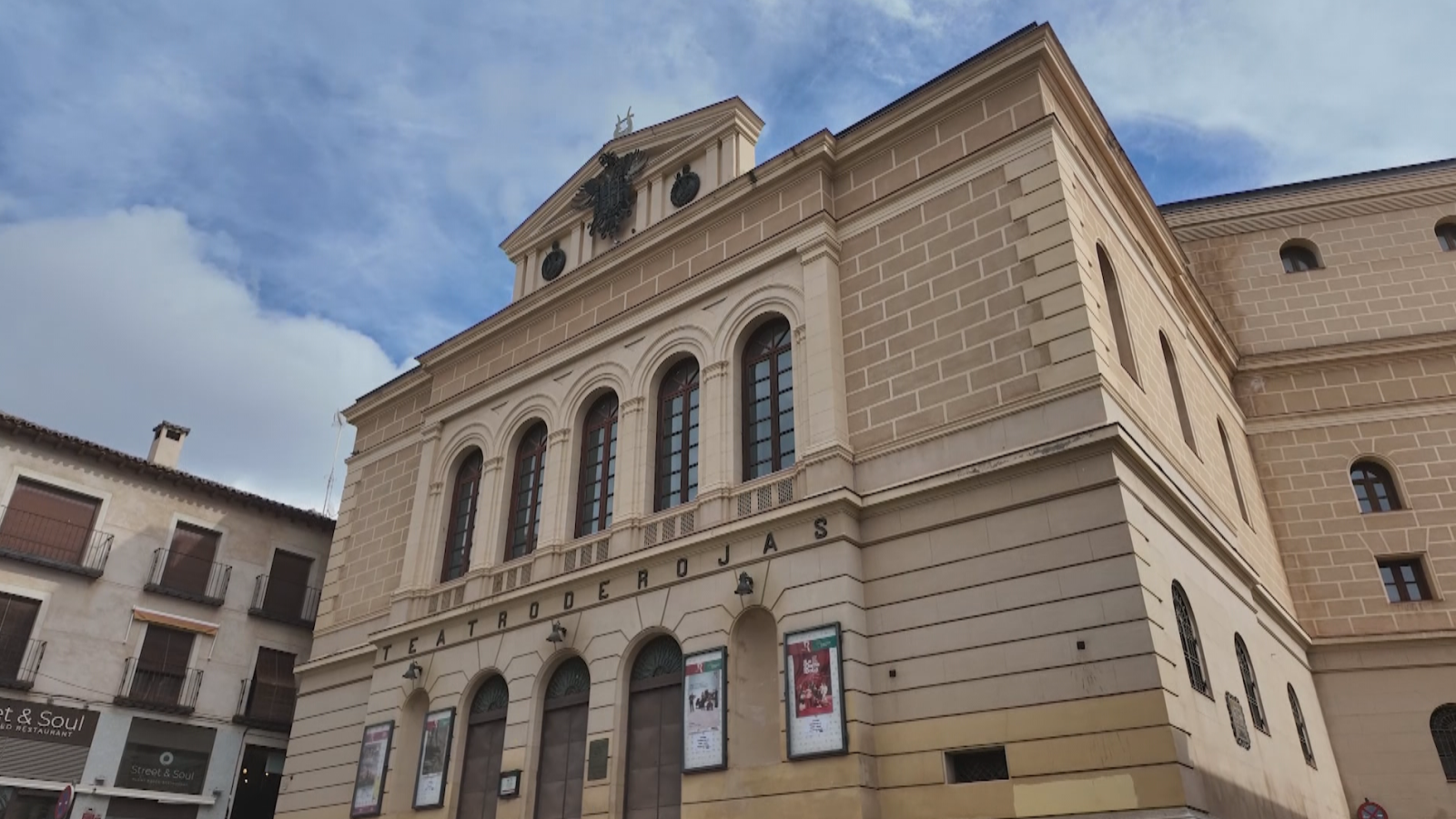 Fachada principal del Teatro de Rojas en Toledo, un edificio decimonónico de tonos ocres con puertas de madera, grandes ventanales acristalados y el escudo imperial en el frontón.