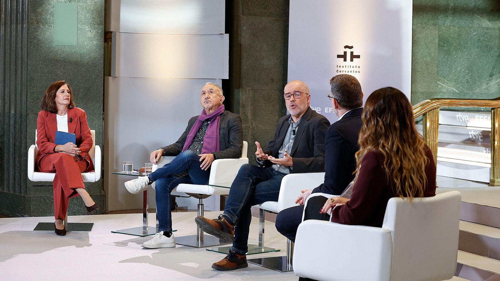Los líderes sindicales Pepe Álvarez y Unai Sordo participan sentados en un evento institucional junto a mesitas de cristal y paneles con logotipos de medios y entidades.