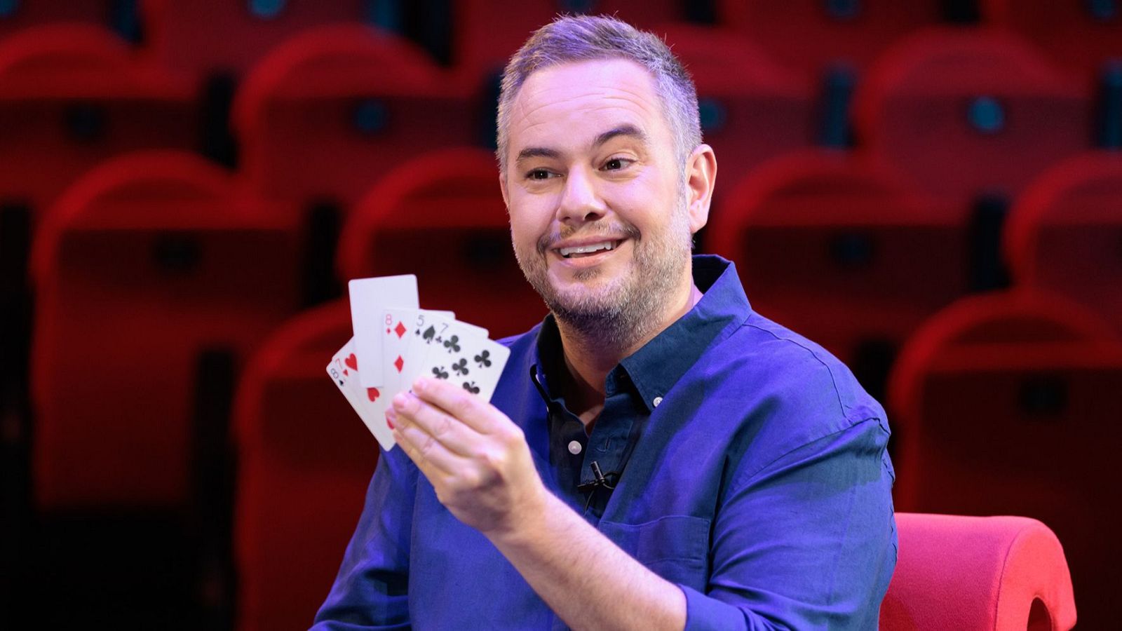 El mago Jorge Blass sonríe amablemente mostrando un abanico de cartas en un teatro con butacas rojas de fondo.