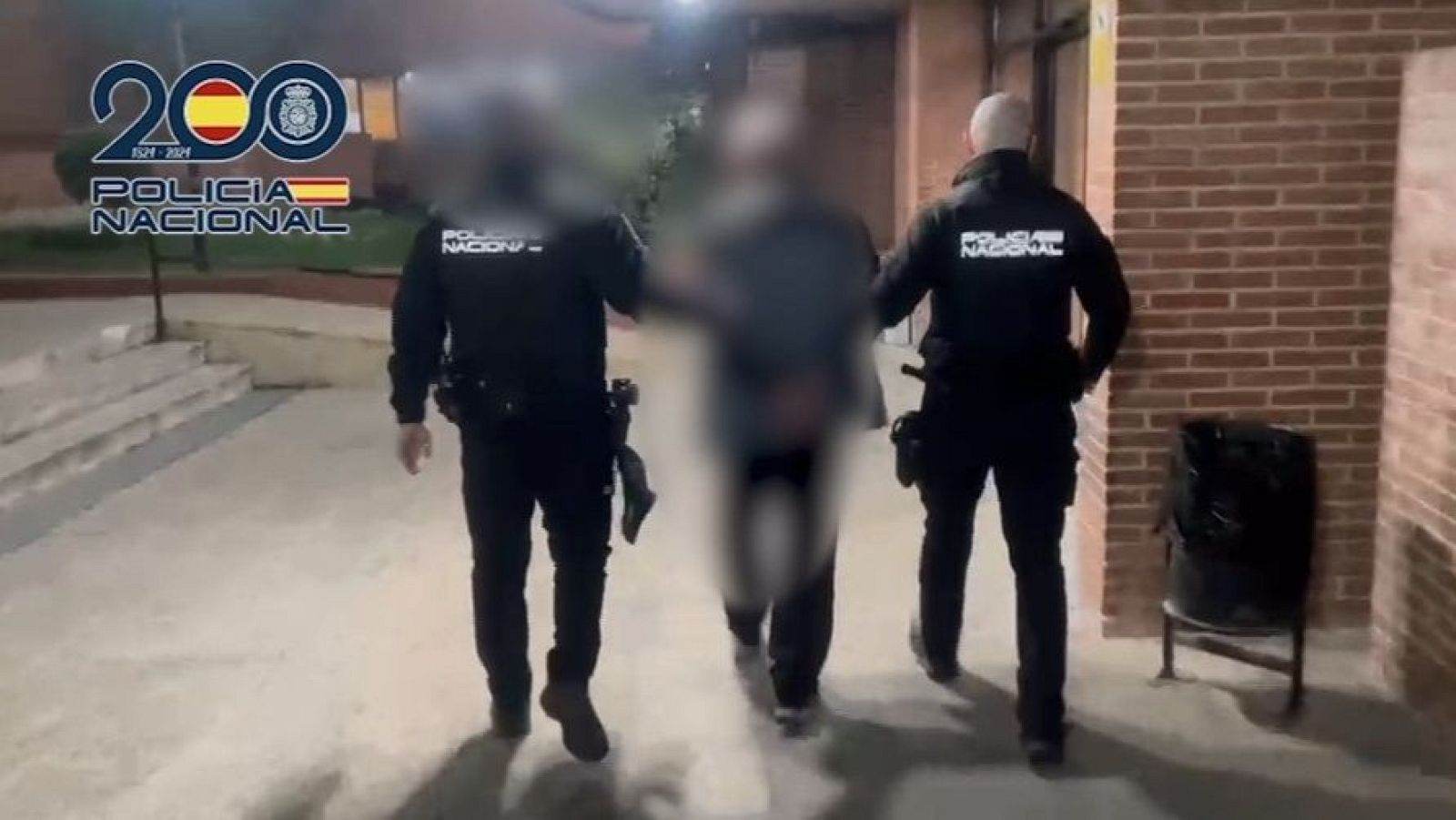 Dos agentes de la Policía Nacional custodian a un detenido durante su traslado nocturno en una zona exterior.
