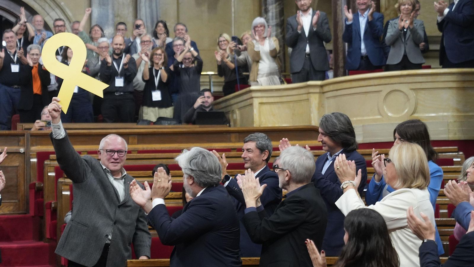 L'exconseller Lluís Puig aixeca un llaç groc al seu escó del Parlament de Catalunya, envoltat de diputats que l'aplaudeixen.