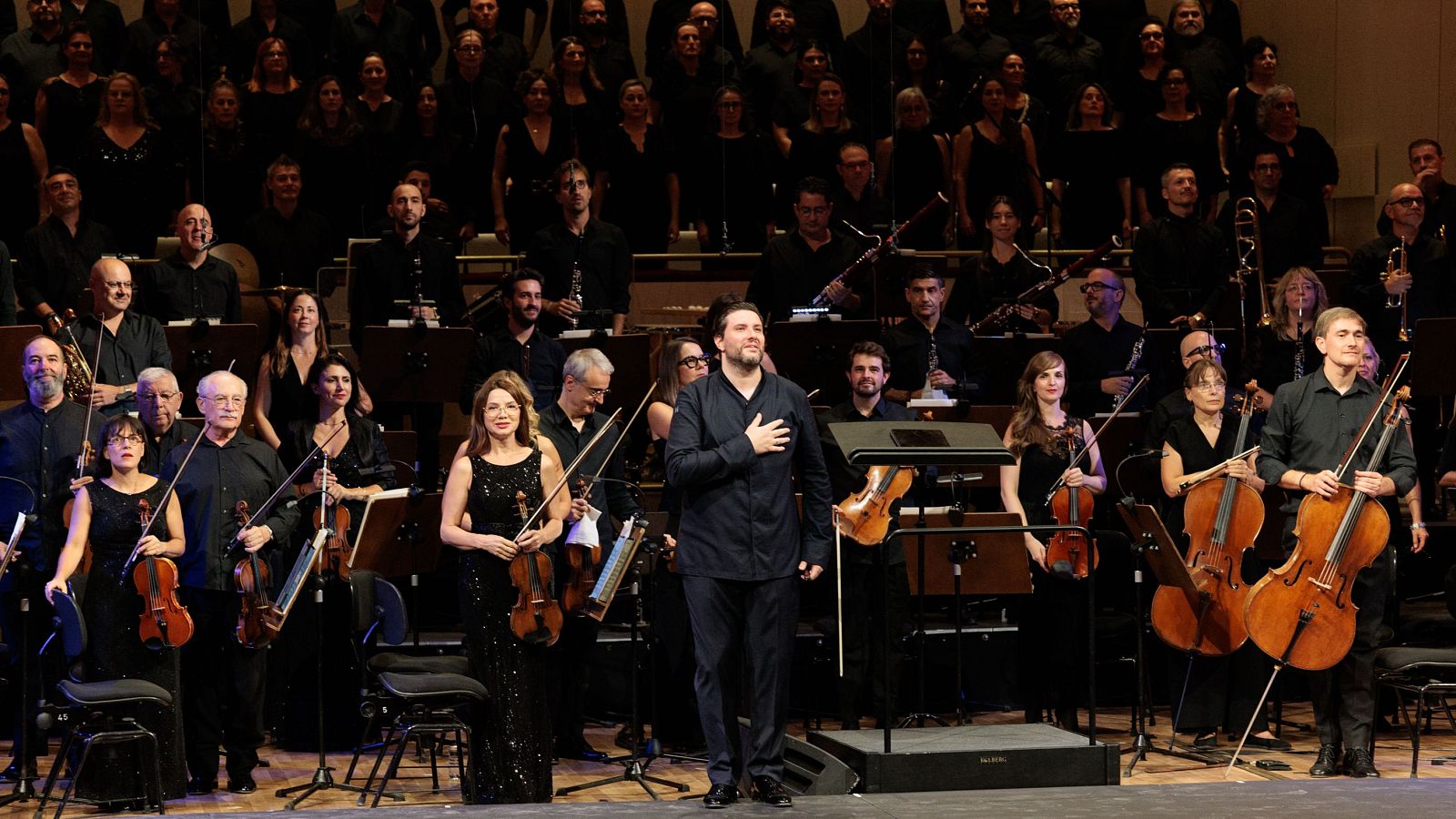 Director de orquesta, la concertino Mariana Todorova con violín y músicos vestidos de negro en el escenario junto al Coro RTVE durante el saludo final de un concierto.