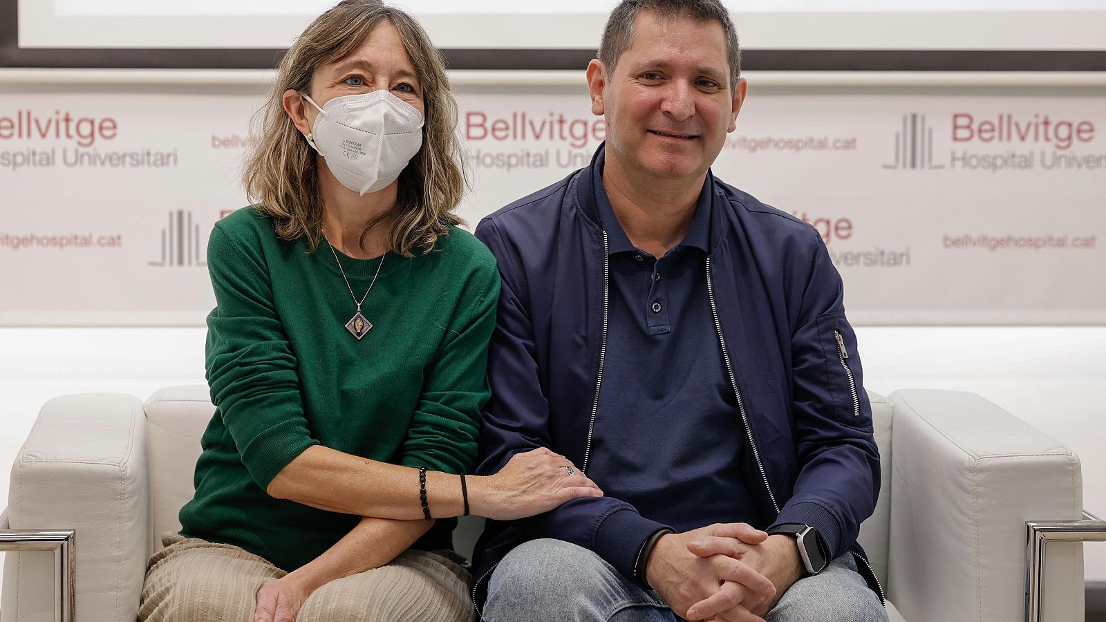 Una mujer con mascarilla y jersey verde apoya su mano sobre el brazo de su marido en un sofá blanco frente al fotocall del Hospital Universitari de Bellvitge