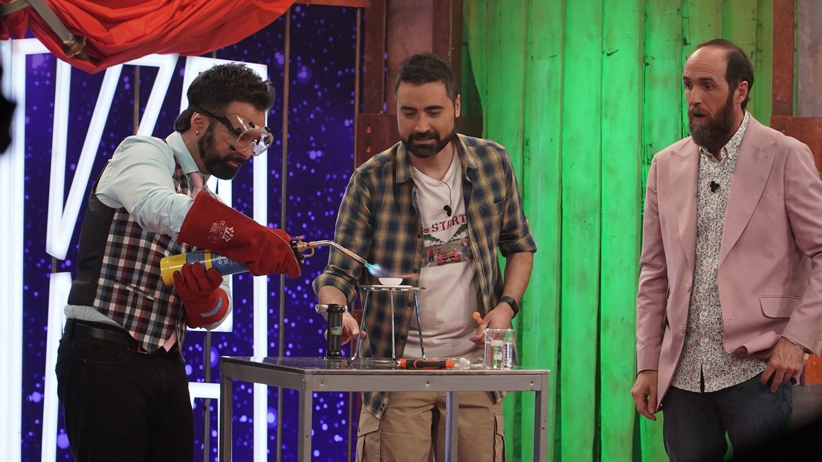 Demostración científica en el plató de un programa de televisión donde tres hombres realizan un experimento con soplete y material de laboratorio ante una mesa de trabajo.