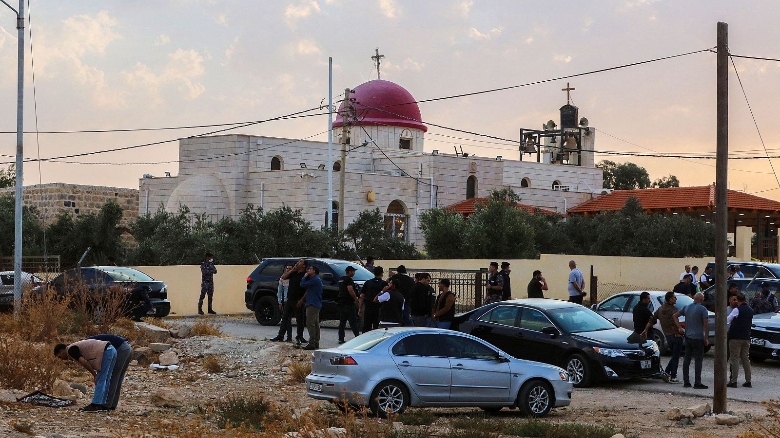 Exterior de una iglesia al atardecer con cúpula roja, rodeada por vehículos estacionados, civiles y fuerzas de seguridad custodiando el perímetro.