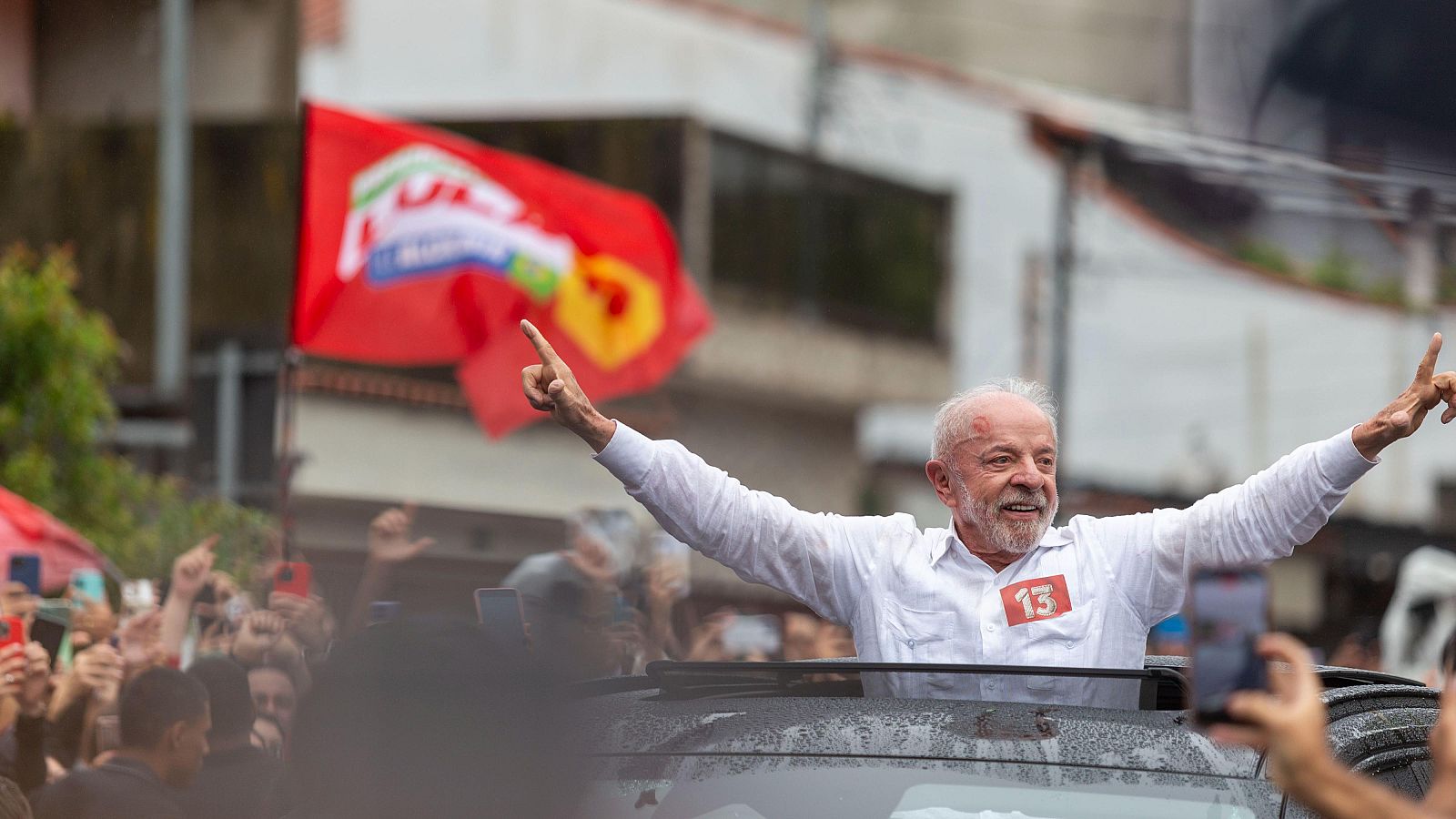 Luiz Inácio Lula da Silva sonríe y extiende los brazos asomado al techo solar de un coche, rodeado de simpatizantes con teléfonos móviles y una bandera roja al fondo.