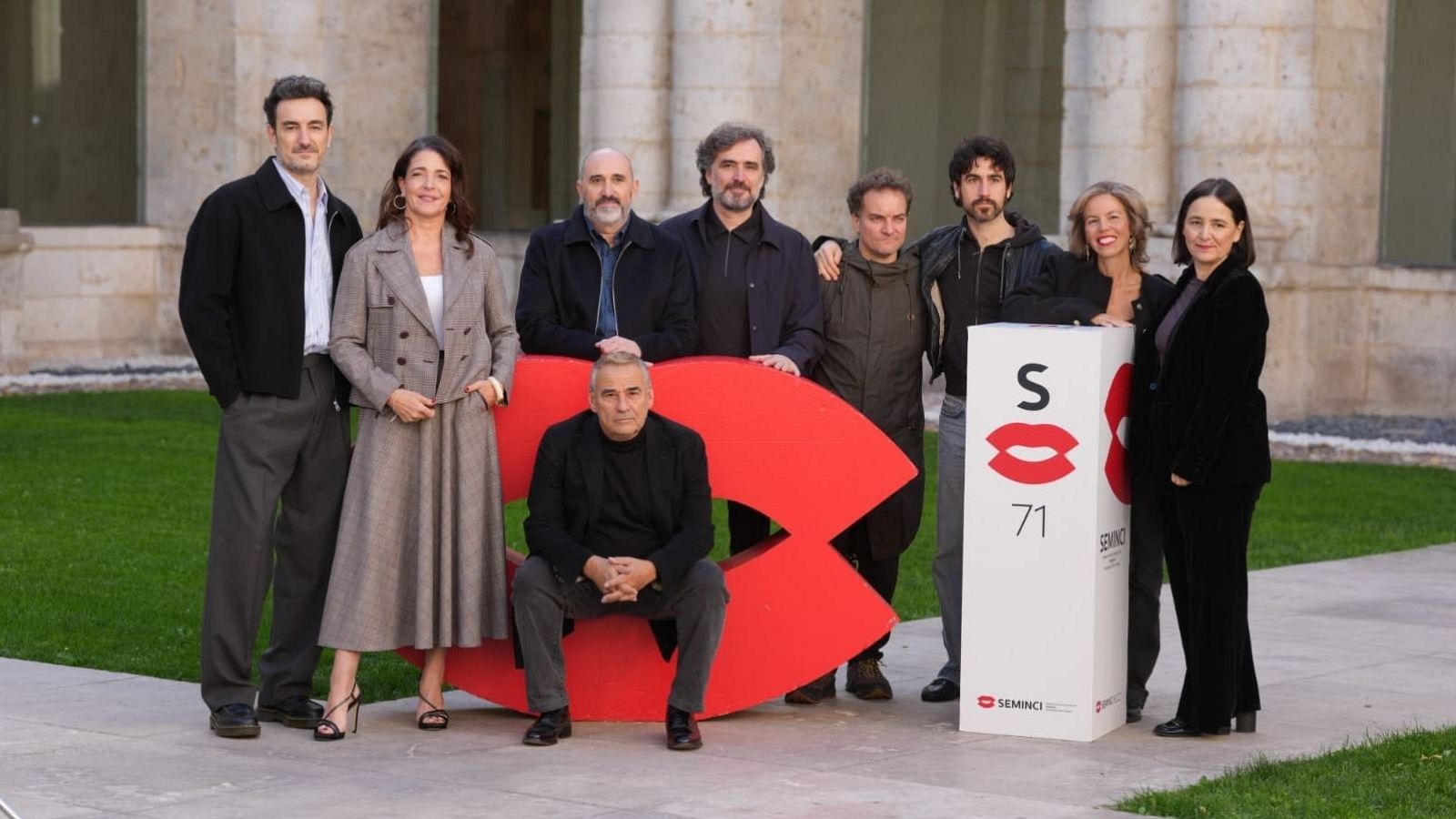 Grupo de nueve personas posando sonrientes junto a una gran escultura roja de labios y un tótem blanco en un claustro histórico.