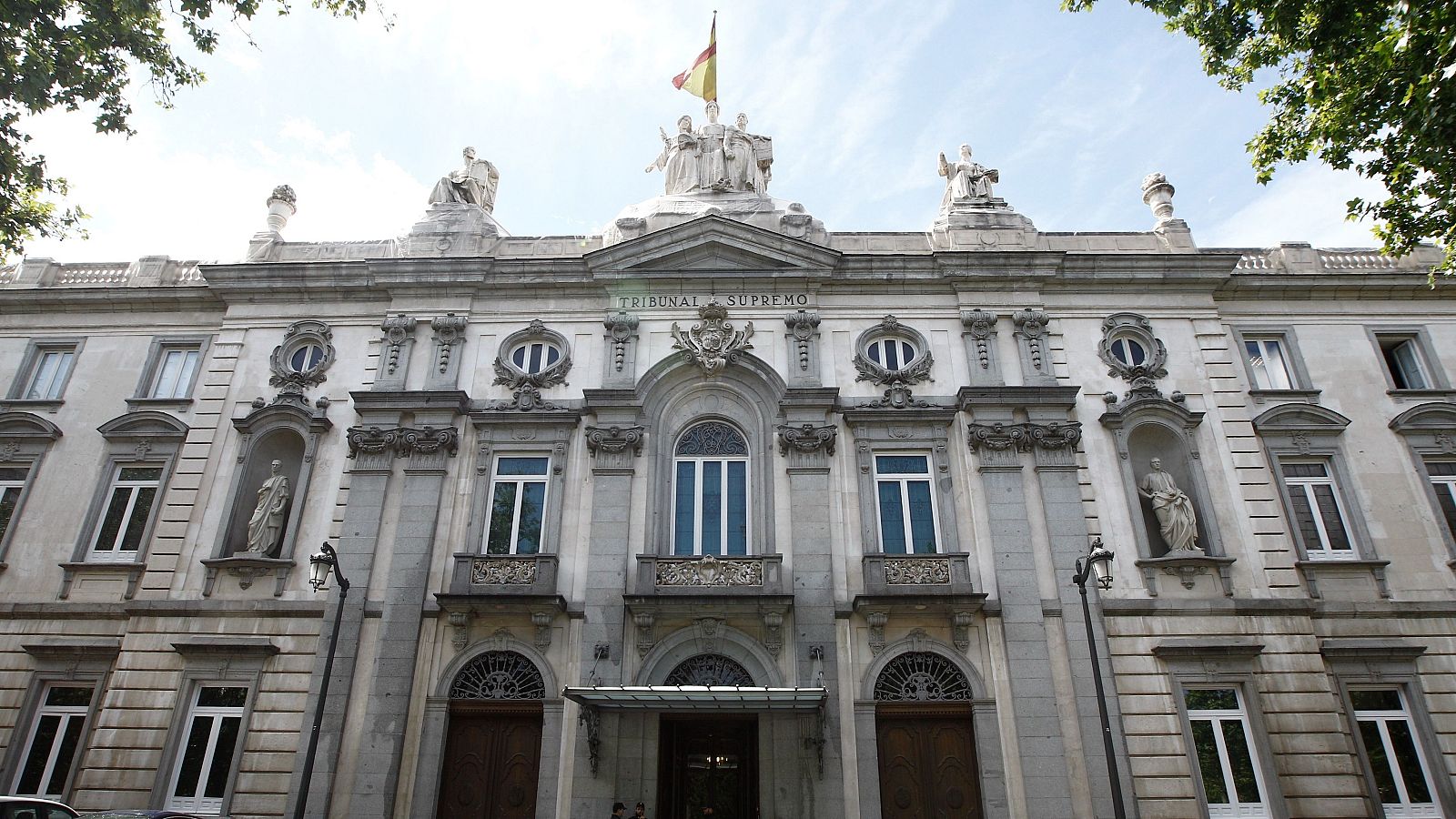 Plano contrapicado de la fachada clásica de piedra del Tribunal Supremo en Madrid, con inscripciones, esculturas, balcones y una bandera de España.