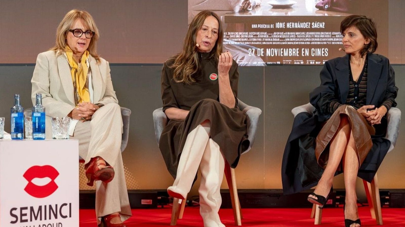 Tres mujeres sentadas en sillones durante una presentación cinematográfica sobre un escenario con moqueta roja y una peana con el logotipo de la Seminci.