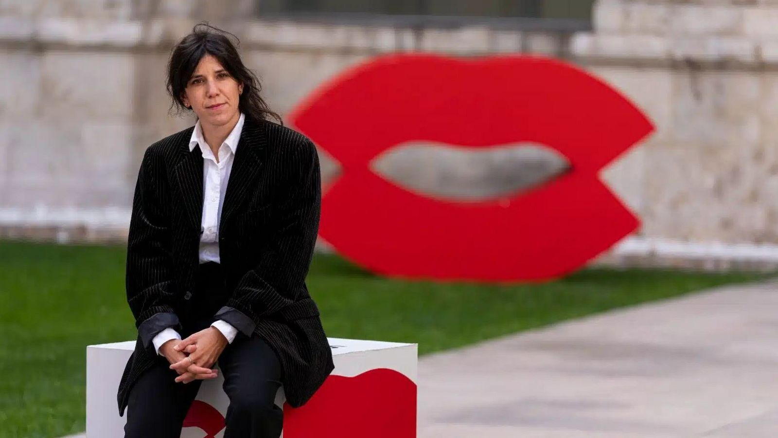 La cineasta Marine Atlan posa sentada en un pedestal blanco con chaqueta oscura, frente a una escultura roja de unos labios y un edificio histórico.