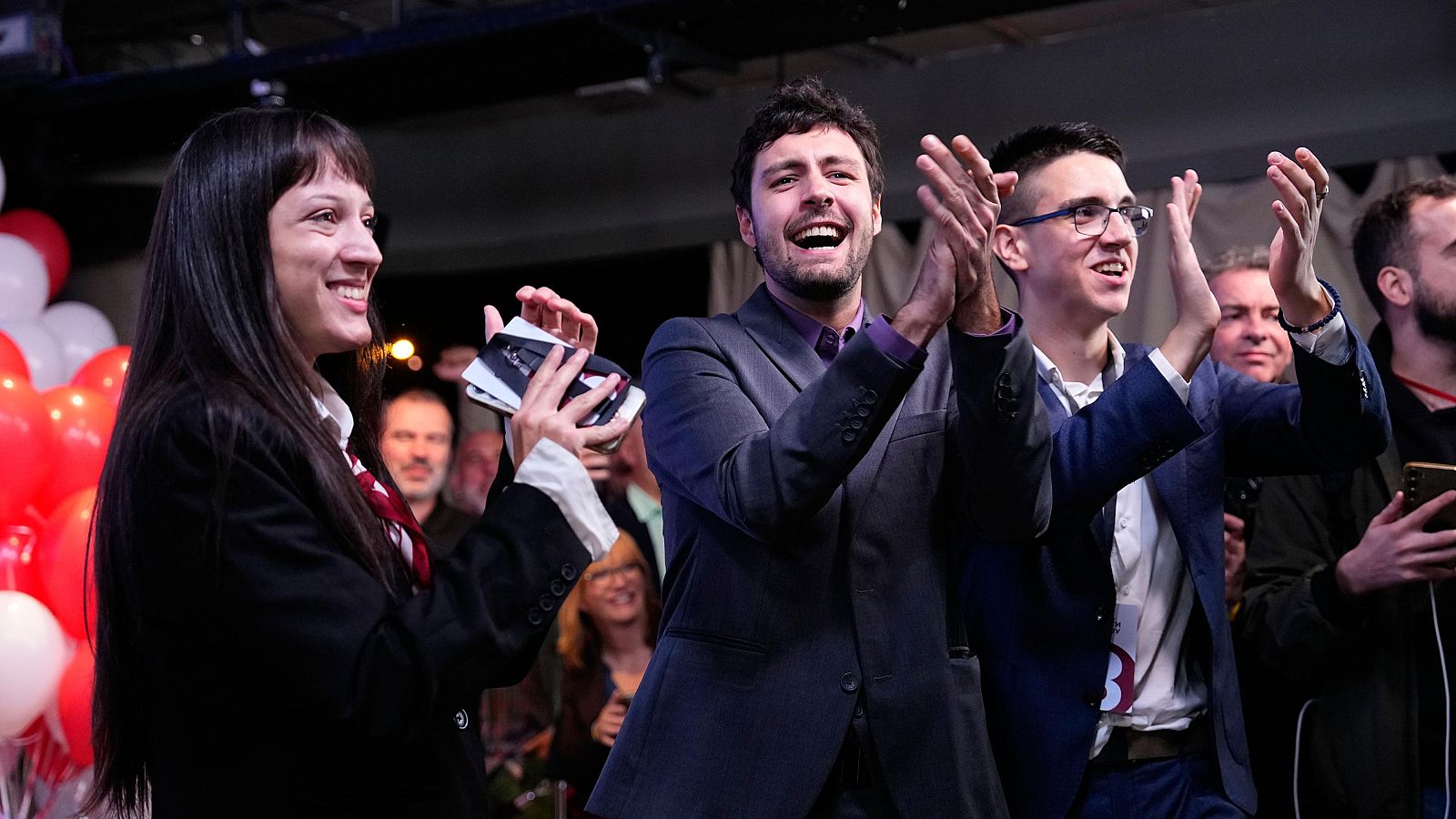 Tres jóvenes representantes estudiantiles celebran eufóricos la victoria electoral sobre un escenario iluminado, aplaudiendo y sonriendo junto a simpatizantes.