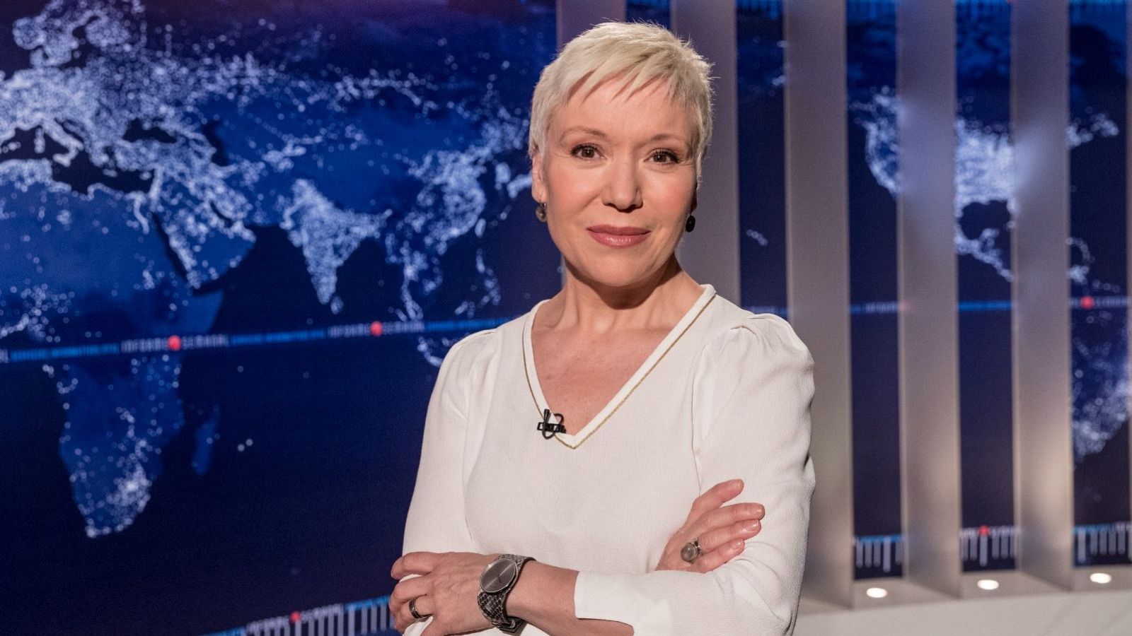 Plano medio de la periodista Rosa Molló en el plató de Informe Semanal, con blusa blanca y brazos cruzados frente a un mapa mundi luminoso.