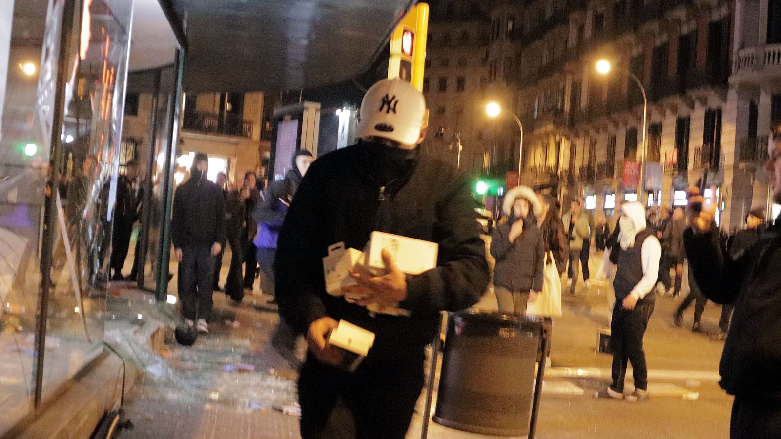 Un jove encaputxat amb una gorra blanca fuig amb caixes de productes robats d'una botiga amb l'aparador destrossat durant una nit de disturbis urbans.