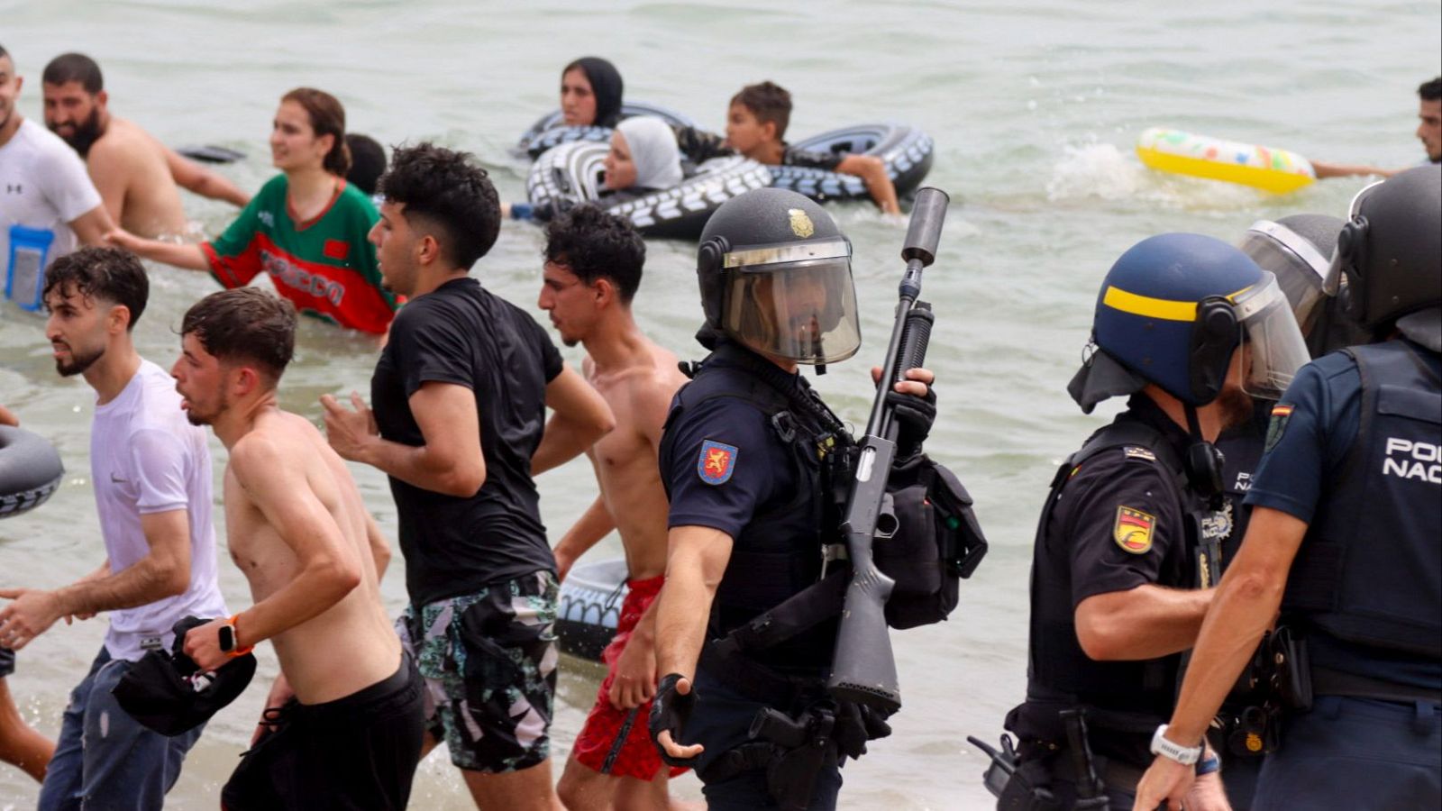 Agentes de la Policía Nacional con cascos y uniformes antidisturbios en el agua, junto a civiles de diversas edades, algunos con flotadores, en una playa. Se observa el escudo de la Policía Nacional y el texto "POLICIA NACIONAL" en los uniformes.