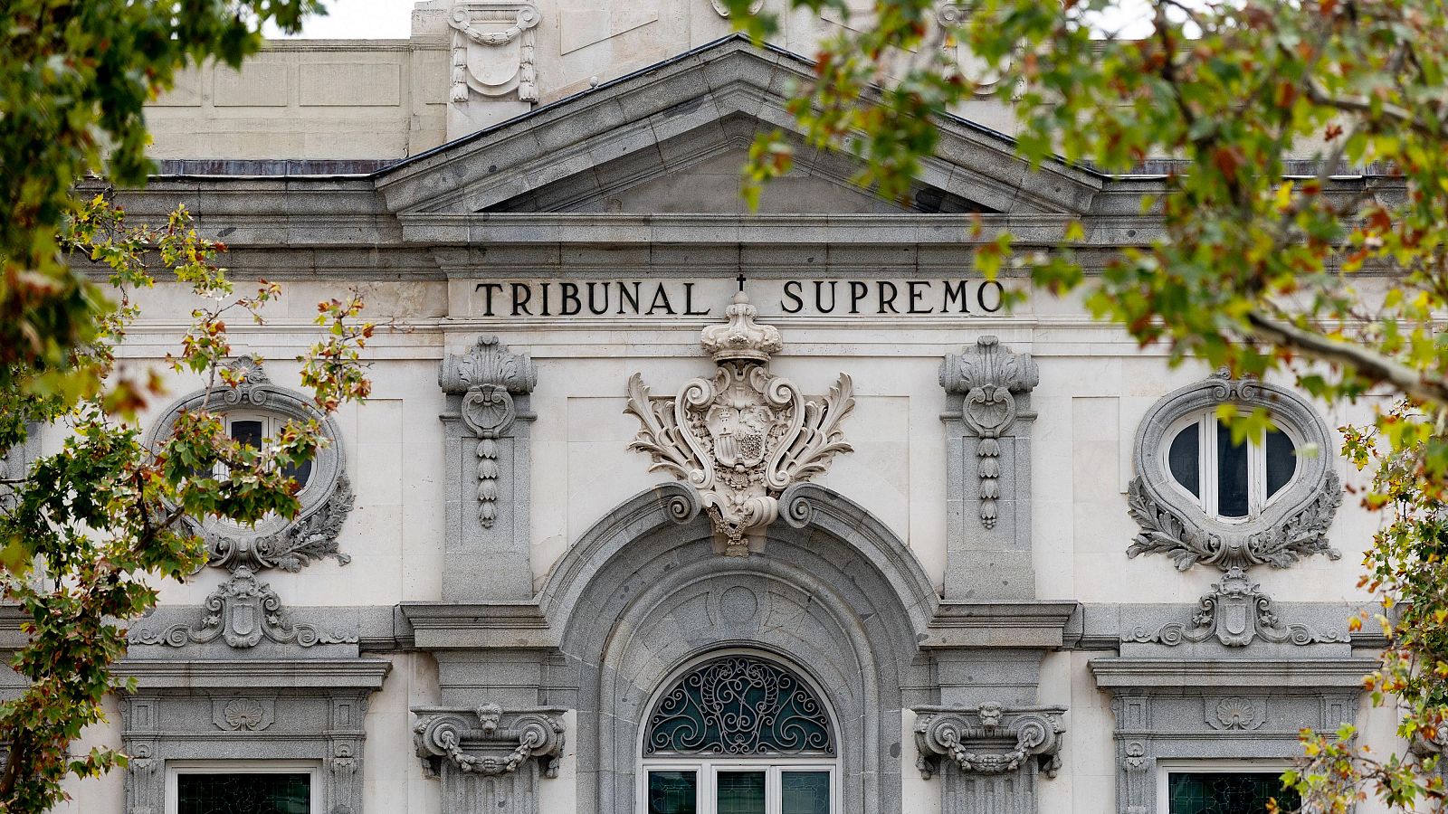 Detalle de la fachada histórica del Tribunal Supremo en Madrid, con su frontón triangular, el letrero en piedra y óculos barrocos, enmarcado por ramas otoñales.
