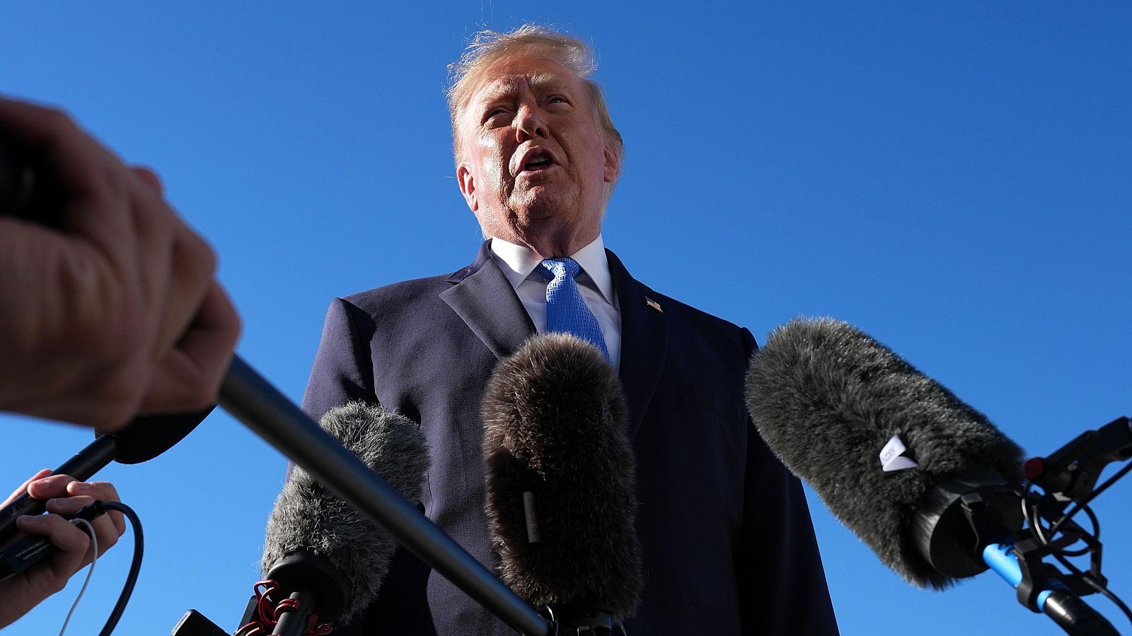Donald Trump habla ante los medios de comunicación bajo un cielo azul, con micrófonos de prensa en primer plano.
