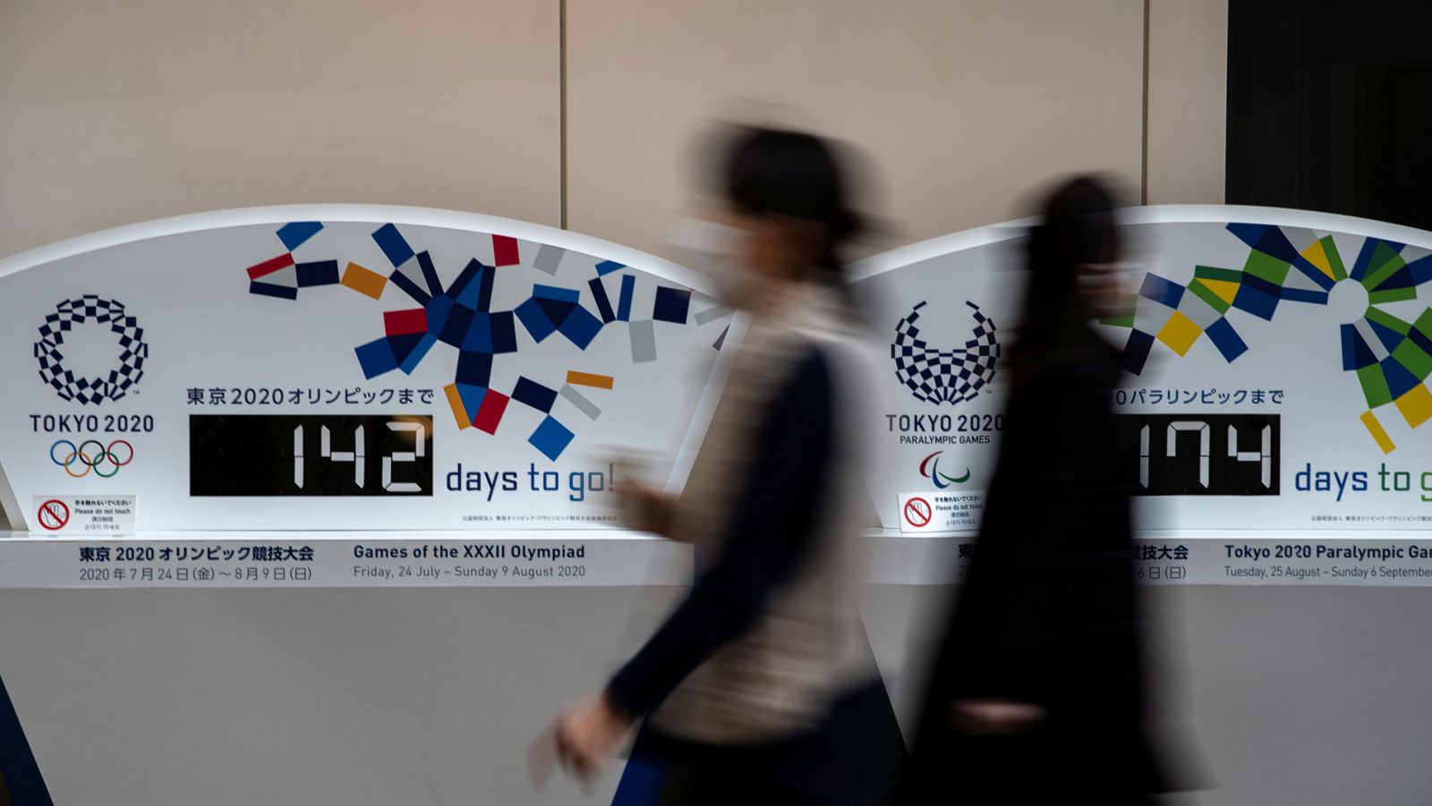 Women wearing protective face masks, following an outbreak of the coronavirus, walk past countdown clocks for the opening ceremony of the Tokyo 2020 Olympic and The Tokyo Paralympic 2020 at a building in Tokyo