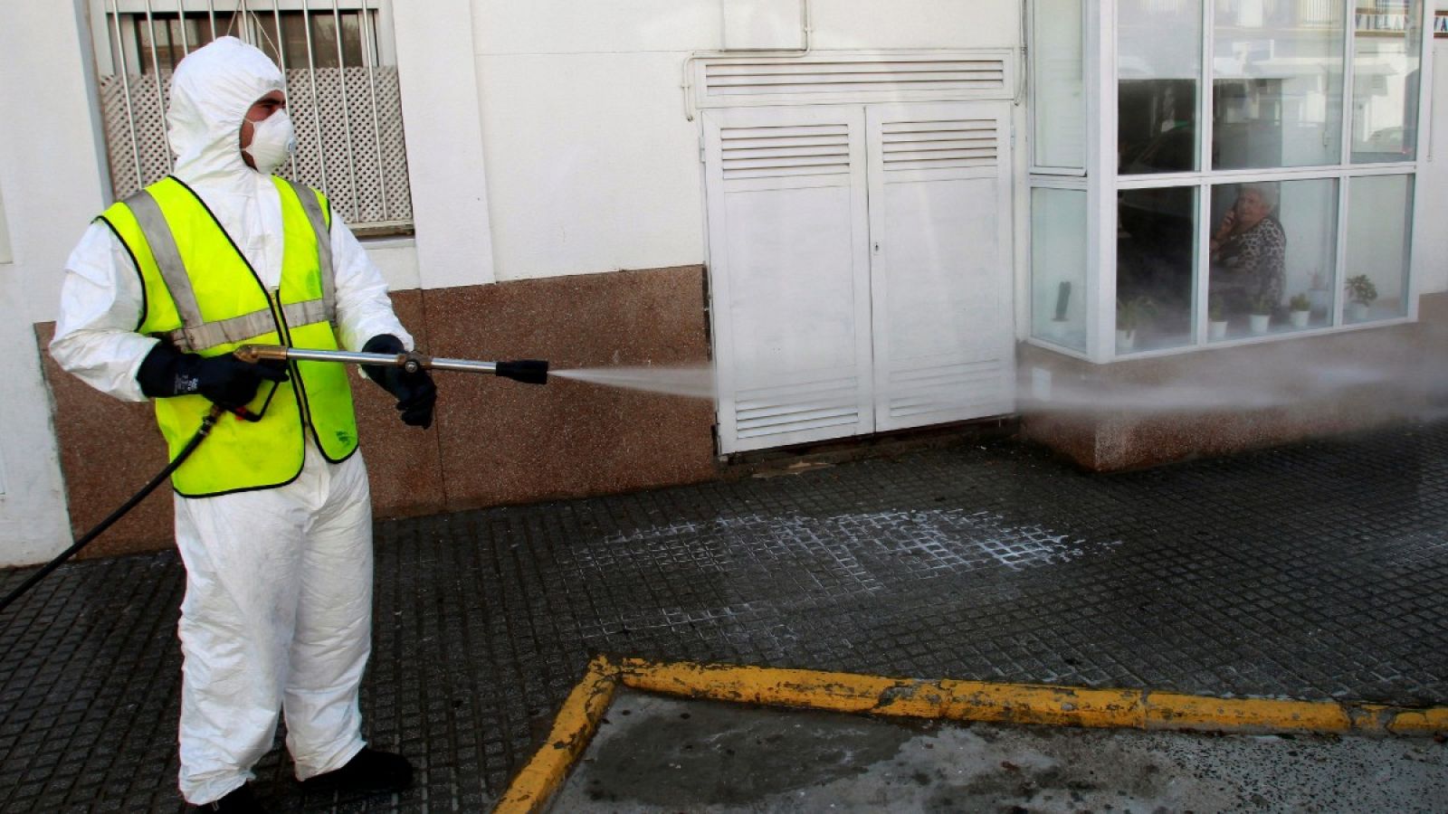 Una mujer habla por teléfono confinada en su casa, mientras observa a un operario desinfectando la calle