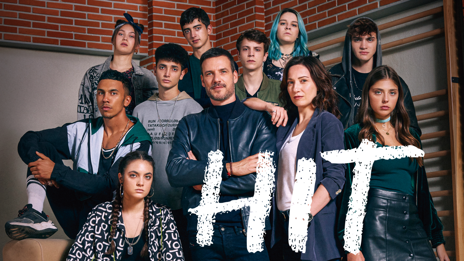 Elenco de "HIT" posando en gimnasio escolar con pared de ladrillo y espaldera. Grupo de jóvenes y dos adultos (hombre con chaqueta negra, mujer con chaqueta azul).