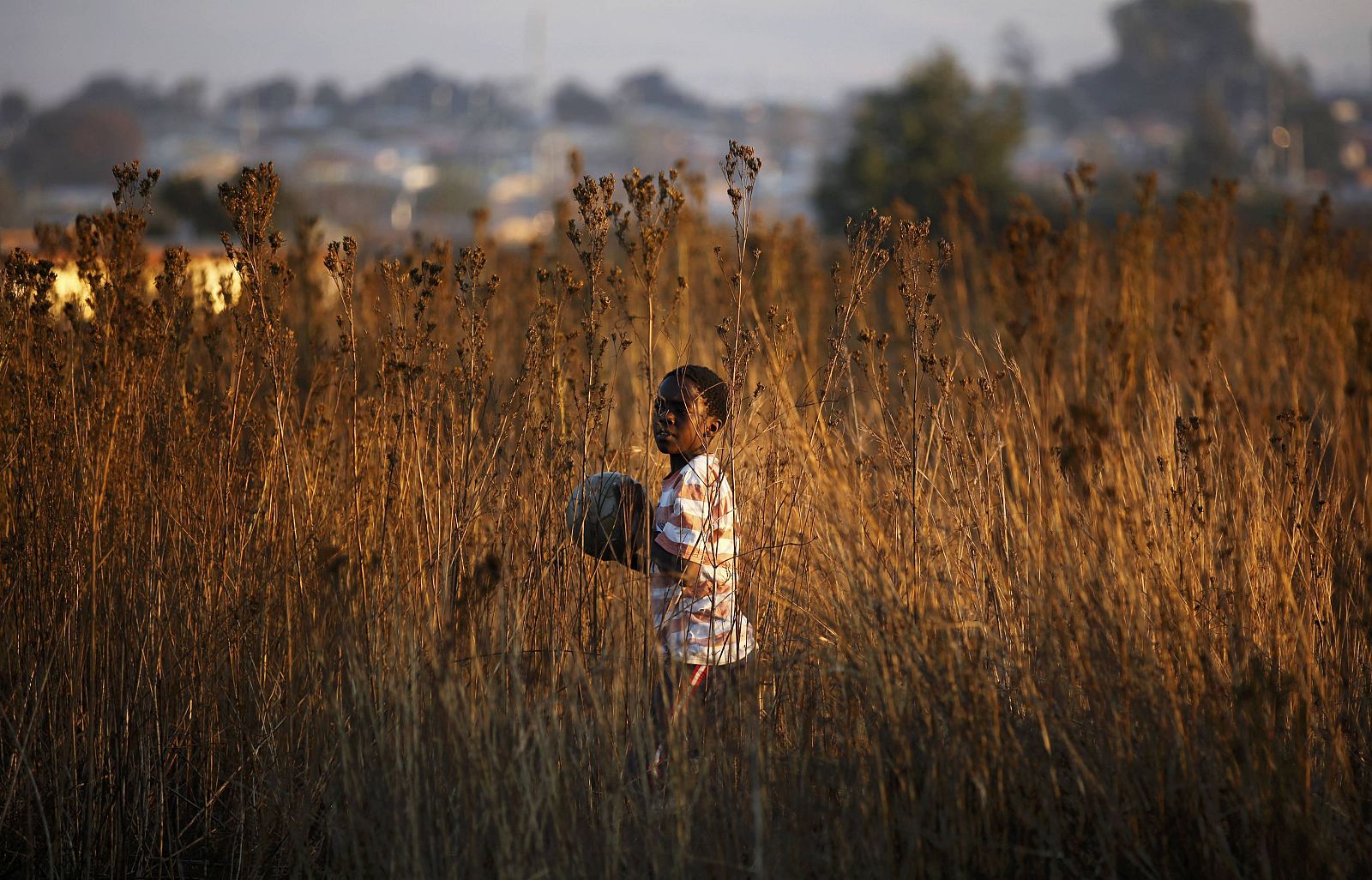 Local child carries a ball while playing soccer at a dirt field in Soweto