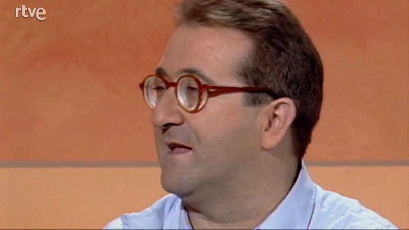 Primer plano de una persona con gafas redondas rojas y camisa azul, hablando posiblemente en una entrevista o presentacin. Fondo salmn, logo RTVE en esquina superior izquierda.