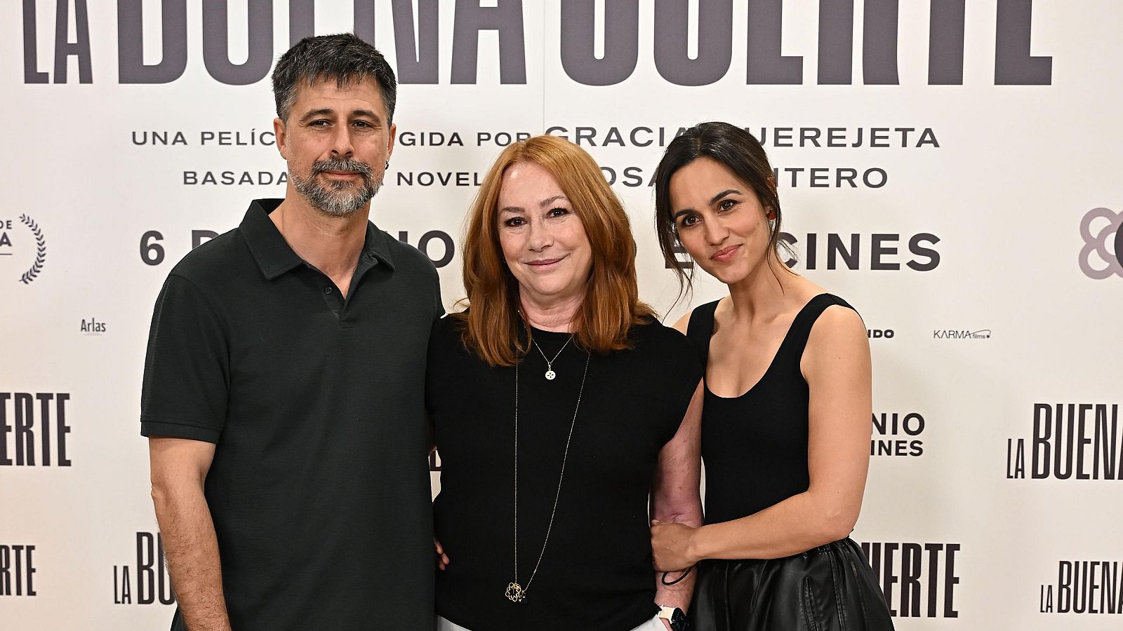 Presentación de la película "La buena suerte": Hugo Silva, Gracia Querejeta y Megan Montaner posan juntos, con atuendos oscuros, ante un cartel promocional.