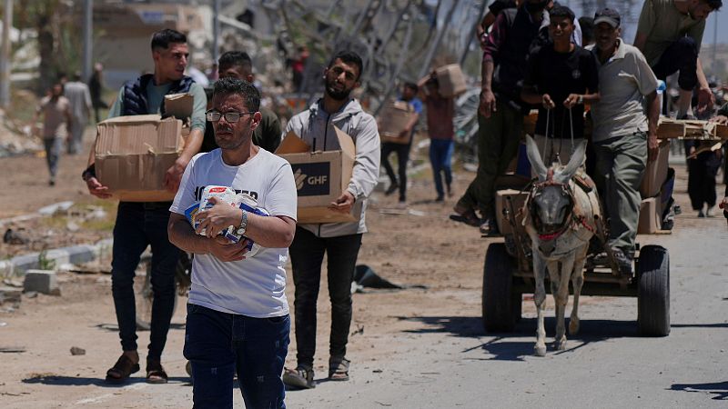 Personas en Gaza transportan ayuda humanitaria, incluyendo alimentos y cajas, en un contexto de edificios dañados. Un burro ayuda al transporte de suministros.