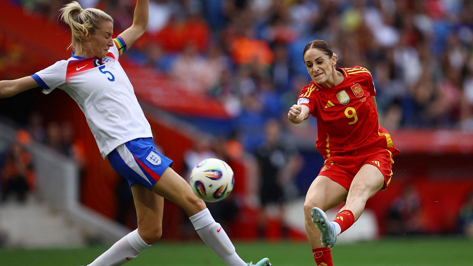 Partido de fútbol entre España e Inglaterra. Jugadora española (roja, #9) y jugadora inglesa (blanca, #5) compiten por el balón en un estadio abarrotado.