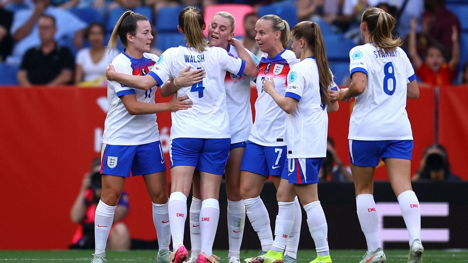 Celebración de jugadoras inglesas de fútbol femenino en un campo, con camisetas blancas y azules, tras un gol o victoria. Se abrazan con alegría y se ven las gradas del estadio.
