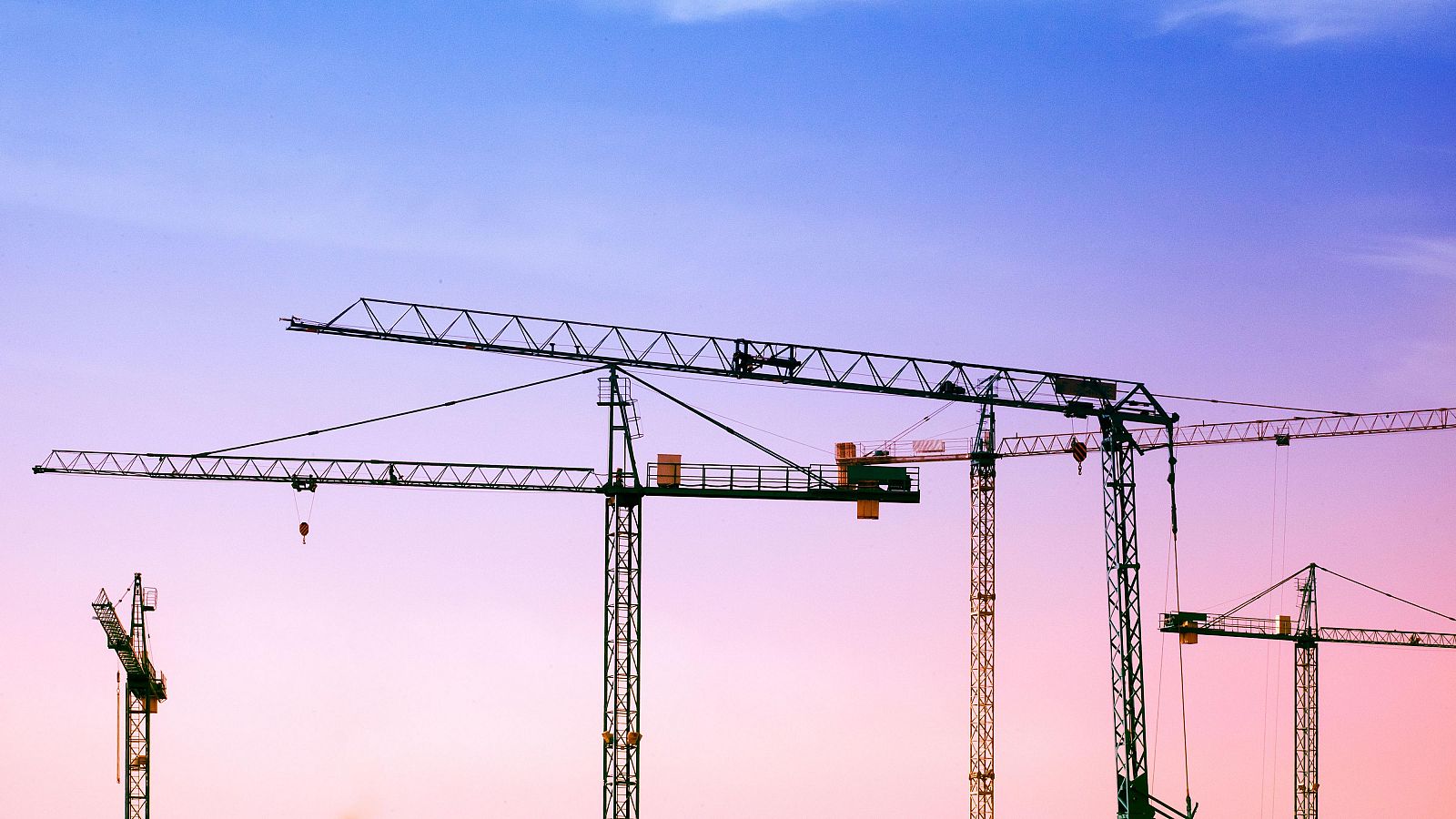 Grúas de construcción de diversos tamaños contra un cielo rosa y morado. La imagen representa la actividad del sector y la construcción de viviendas.