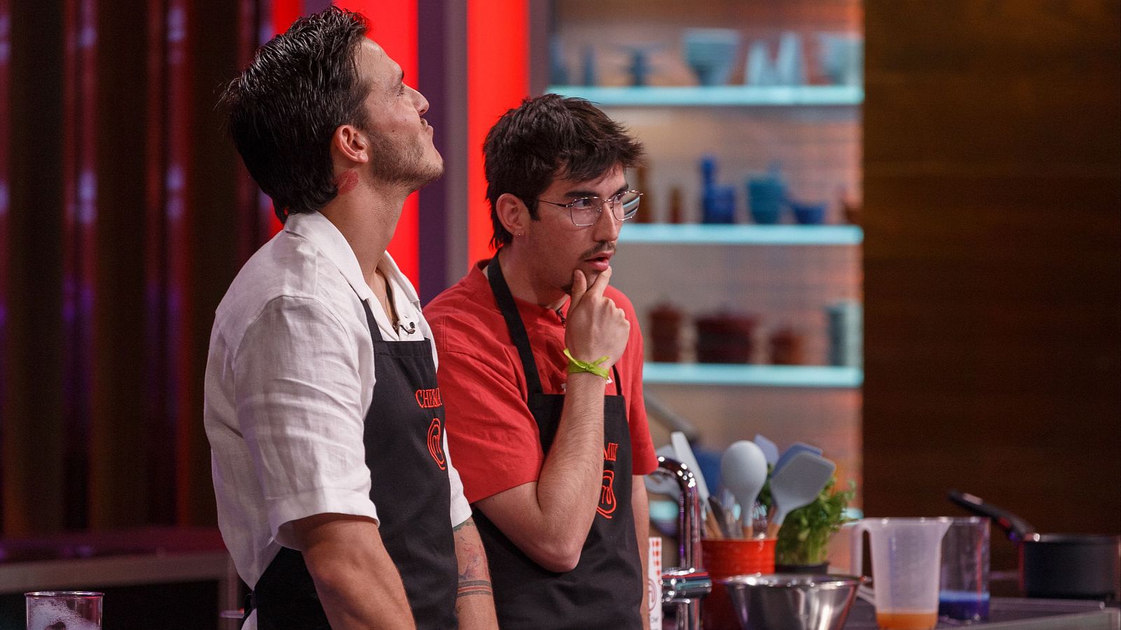 En la cocina de MasterChef, dos concursantes, uno con camisa blanca y otro con camiseta roja y gafas, reaccionan a un reto de eliminación; uno sorprendido, el otro pensativo.