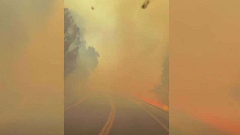Carretera con llamas a un lado, envuelta en denso humo anaranjado y amarillento. Visibilidad reducida por incendio activo.