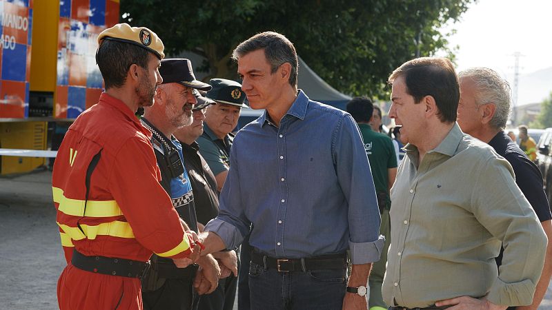 Pedro Sánchez, con camisa vaquera, estrecha la mano a un miembro de emergencias con uniforme rojo y boina. Al fondo, personal de seguridad y un vehículo de "MANDO".