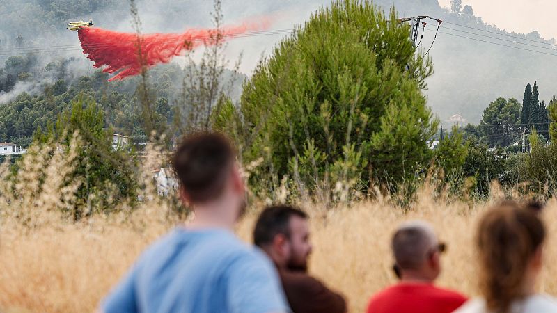 Avión amarillo descarga retardante rojo sobre incendio forestal con casas y montañas. Cuatro personas observan desde un campo de hierba seca.