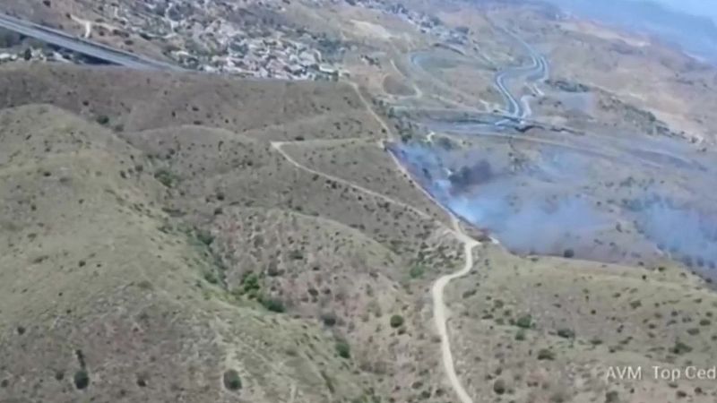 Paisaje montañoso seco con humo de incendio, caminos de tierra y una carretera en la distancia. Texto: 'AVM Top Ced'.