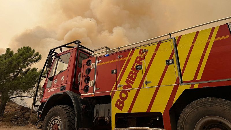 Camión de bomberos "BOMBERS CPBC 431" rojo con franjas amarillas, ante un cielo cubierto de humo ocre y anaranjado. Se aprecian un árbol y suelo árido.