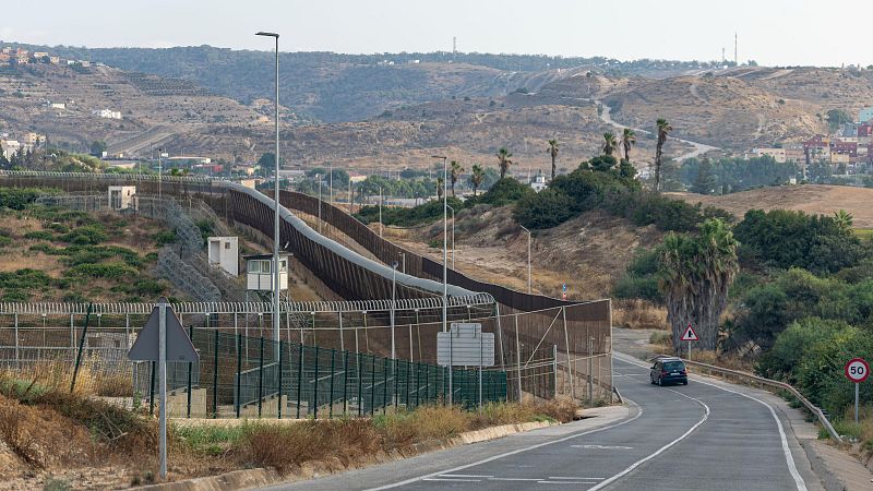 Vista de la valla fronteriza de Melilla con carretera, coche, señales de tráfico, garitas de vigilancia y concertinas sobre terreno árido.