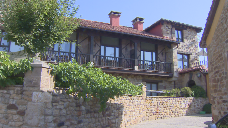 Edificio rural "Alojamientos Eclipse" en Valderredible, Cantabria. Fachada de piedra, tejado rojizo, balcón de madera y vegetación circundante bajo un cielo azul.