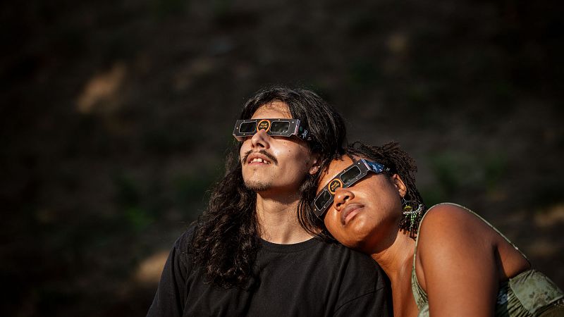 Un hombre y una mujer observan un eclipse solar en Lugo, Galicia, con gafas de protección. Él tiene cabello largo y barba, ella piel oscura y rastas, apoyada en su hombro. Ambos miran al cielo.