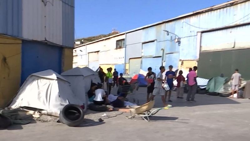 Campamento improvisado con tiendas de campaña y un grupo de personas en un entorno industrial bajo un cielo azul.