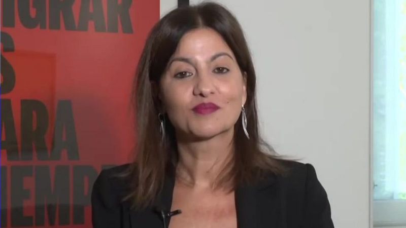 Mónica López, presentadora española, en primer plano con labios fucsia, chaqueta oscura y pendientes plateados. Fondo rojo con texto parcial y pared blanca con luz.